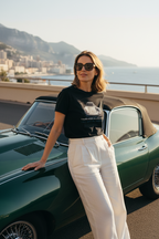  A photorealistic candid shot of a sophisticated woman in her late 30s, wearing the "Timeless Allegiance" tee tucked into high-waisted linen trousers. She is leaning casually against a perfectly restored vintage 1960s Jaguar E-Type convertible, parked on a sun-drenched coastal road in Monaco. Her sunglasses are on, wind blowing slightly through her hair, exuding relaxed luxury. Film grain texture. 