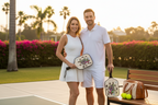 Lifestyle:** A sun-drenched, upscale outdoor pickleball court at a private club during golden hour. A stylish couple in elegant white activewear is smiling, holding the Floral Serpent paddles. One paddle is resting on a teak bench next to an open leather-trimmed carry bag showing the balls. The atmosphere is relaxed luxury. 