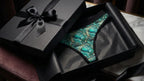 Luxury gift packaging, a black matte box half open, emerald patterned thong inside wrapped in black tissue paper, elegant ribbon, cinematic lighting.