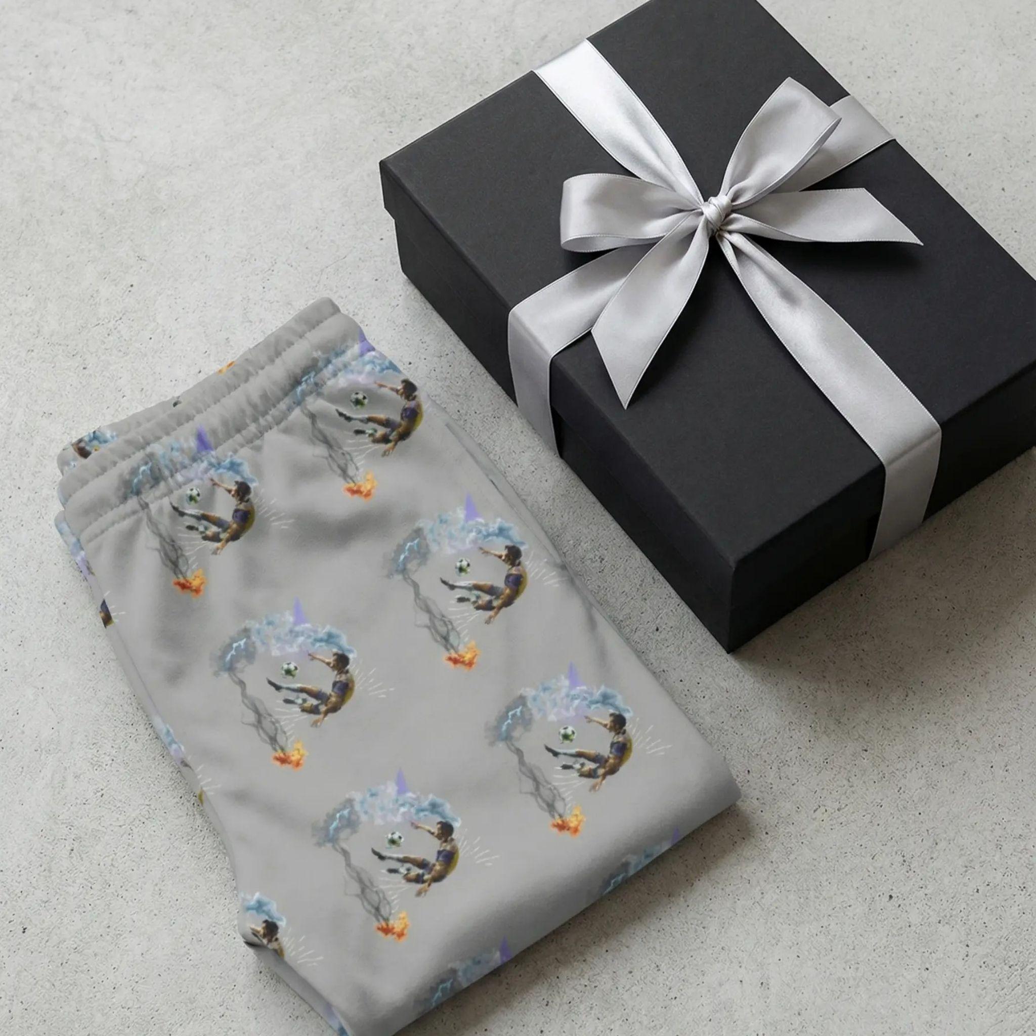 Flat lay photography of the folded joggers next to a premium black gift box with a silver ribbon, minimalist aesthetic, high-end boutique feel.